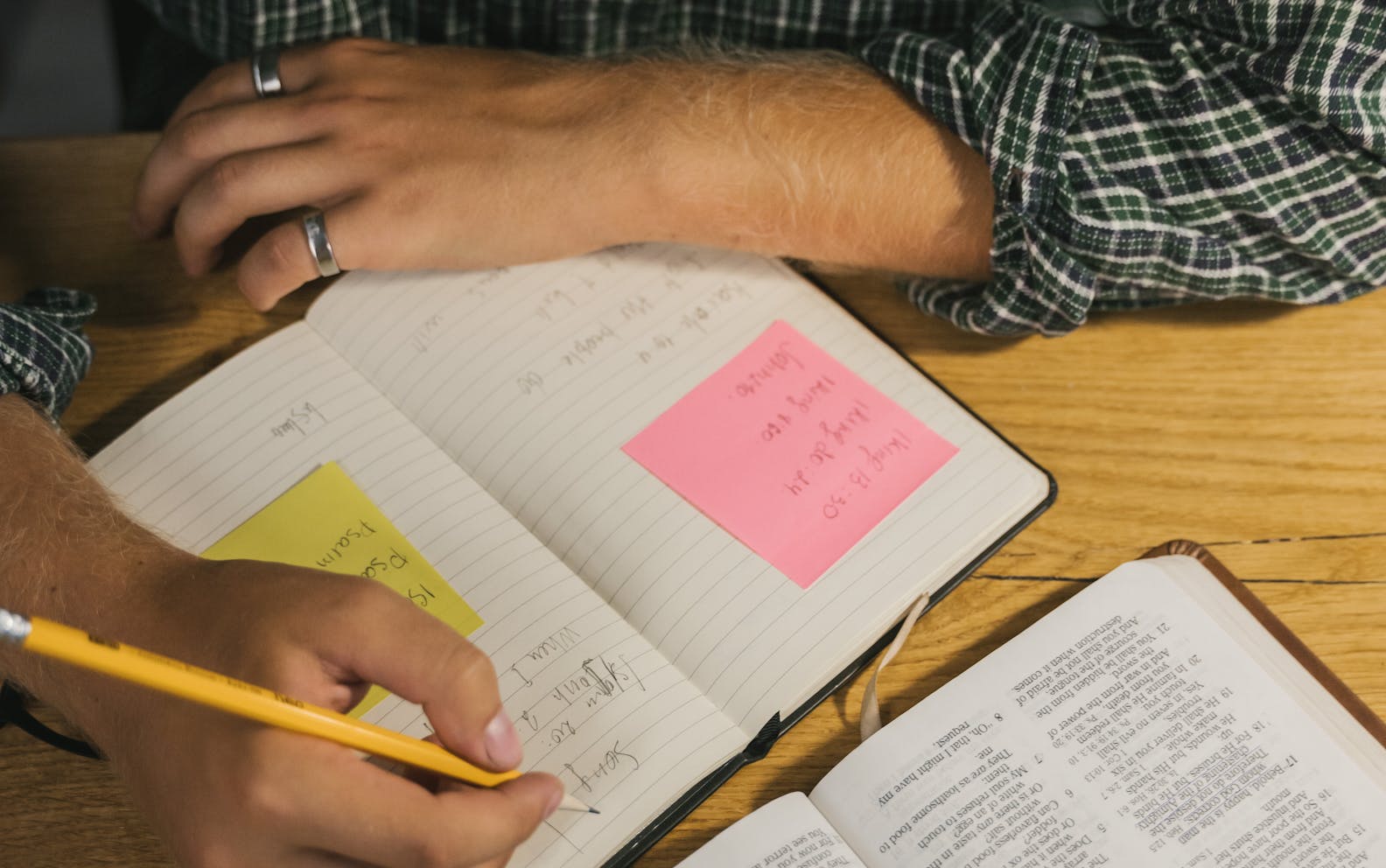 A man writes notes in a notebook while reading an open Bible on a wooden table.